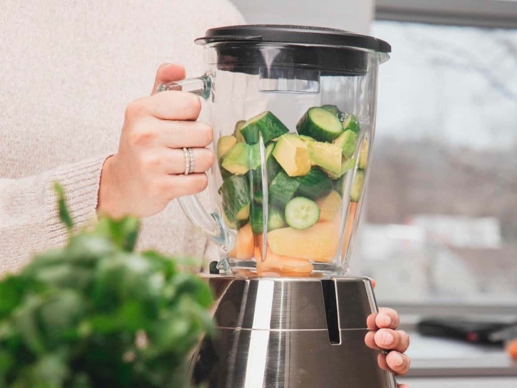 A person holds a blender filled with chopped cucumbers and other fruits or vegetables, preparing to blend them. The scene is set in a bright kitchen with a leafy green plant in the foreground. GREENHOUSE Goodness