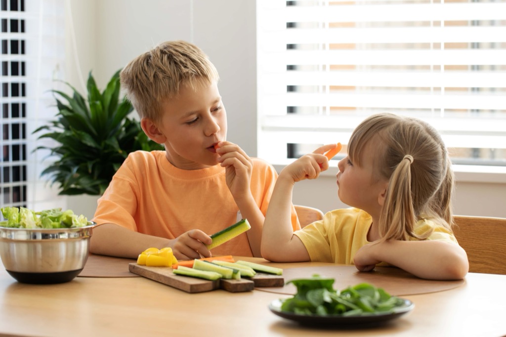 Two young children sit at a table, eating fresh vegetables. The boy holds a cucumber slice, and the girl holds a carrot stick. Sliced vegetables and a metal bowl are on the table, with sunlight streaming through blinds. GREENHOUSE Goodness