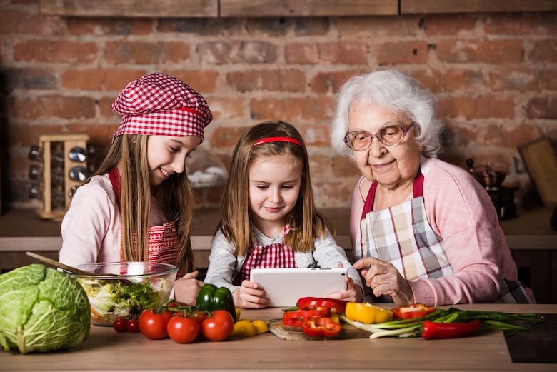An elderly woman and two young girls, all in aprons, sit at a kitchen counter with fresh vegetables. The girls smile at a tablet while preparing food, including bright Tomatoes on the Vine, creating a warm and joyful cooking scene. GREENHOUSE Goodness