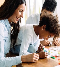 A woman and a young girl smile and draw together at a table, using colorful markers and paper. Another person is partially visible in the background, also engaged in the activity. GREENHOUSE Goodness