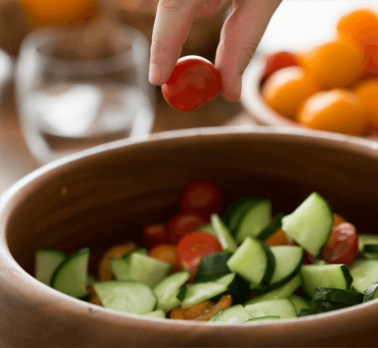 A hand drops a cherry tomato into a wooden bowl filled with sliced cucumbers and tomatoes, with more tomatoes and a glass blurred in the background. GREENHOUSE Goodness