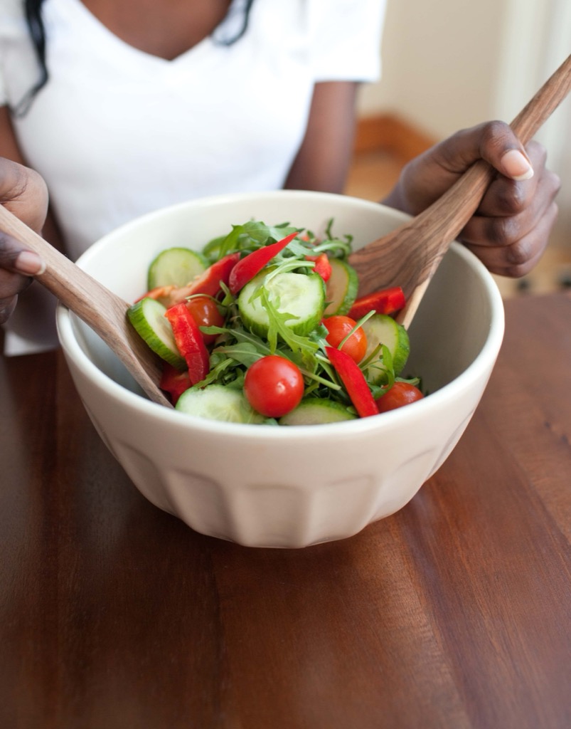 A person in a white shirt is mixing a fresh salad with wooden utensils in a white bowl. The salad contains cucumbers, cherry tomatoes, red bell peppers, and arugula. GREENHOUSE Goodness