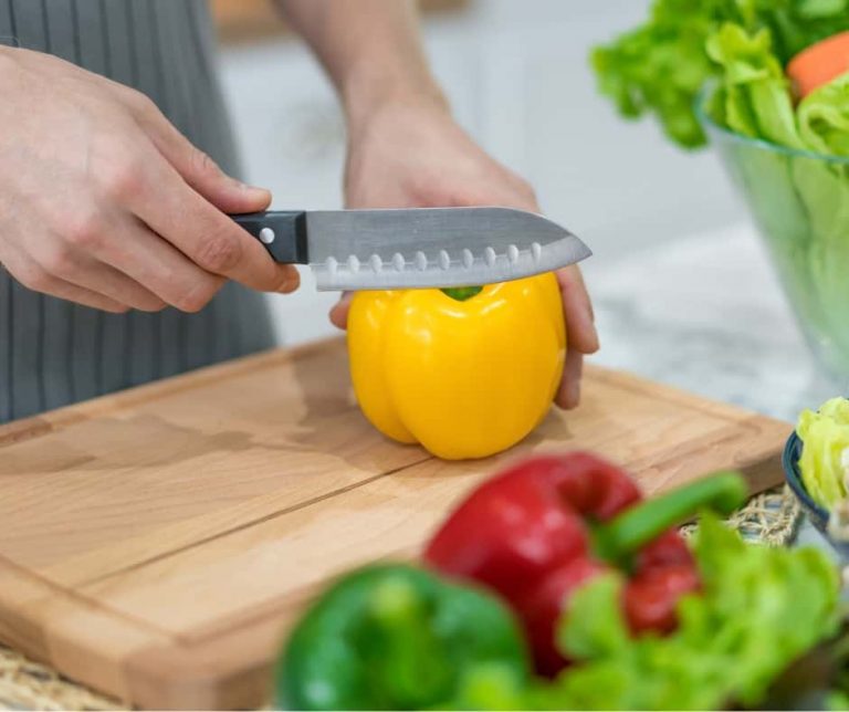 A person slicing a yellow bell pepper on a wooden cutting board, with red and green bell peppers and leafy greens nearby. GREENHOUSE Goodness