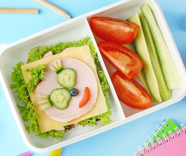 A lunchbox with sliced tomatoes, cucumber sticks, and a sandwich decorated to look like a face using cucumber, blueberry, and a red pepper slice, on a blue background with a notebook and colored pencils nearby. GREENHOUSE Goodness