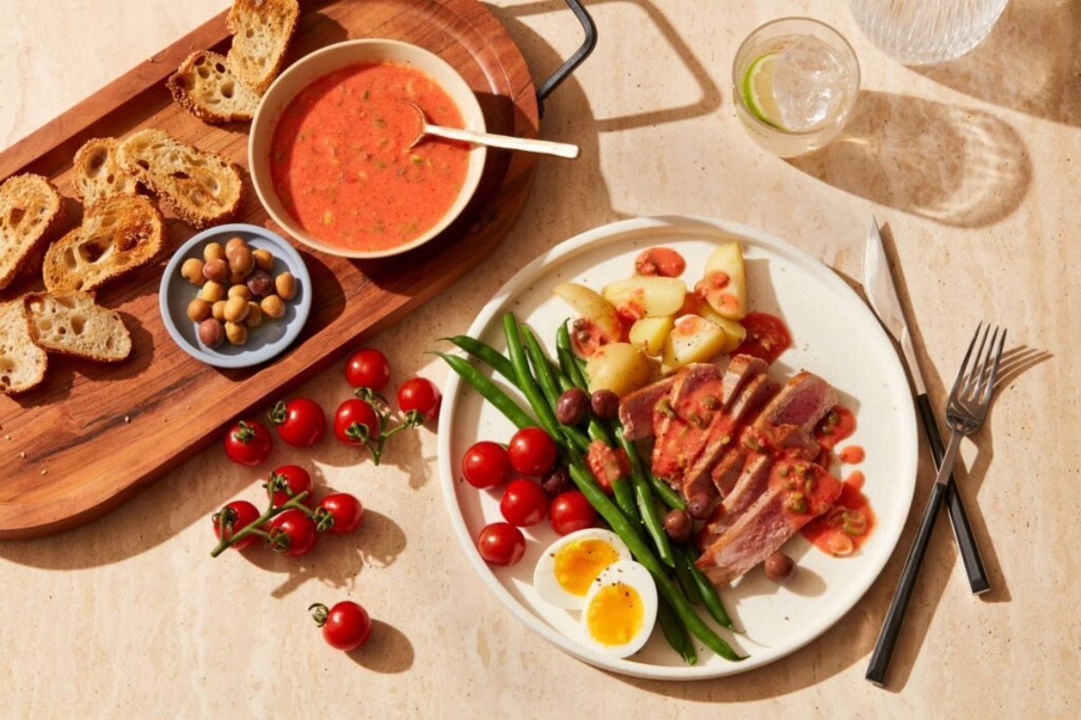 A plate with seared tuna, green beans, cherry tomatoes, potatoes, a halved soft-boiled egg, and olives, next to a bowl of tomato soup, toasted baguette slices, and a small dish of mixed olives on a light table. GREENHOUSE Goodness