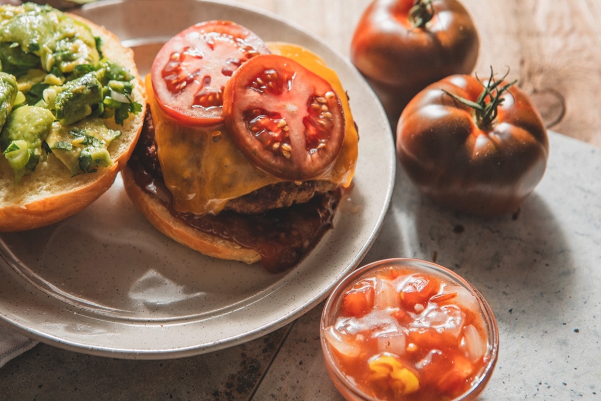 A cheeseburger topped with sliced tomatoes sits open-faced on a plate, with a bun spread with avocado beside it. Nearby are two whole tomatoes and a small bowl of tomato and onion salsa. GREENHOUSE Goodness