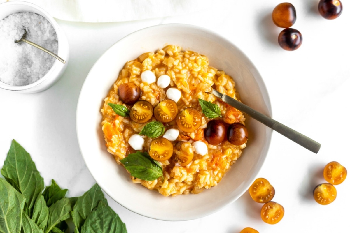 A bowl of risotto topped with yellow cherry tomatoes, small mozzarella balls, brown cherry tomatoes, and fresh basil leaves. A spoon rests in the bowl, with salt and more tomatoes on the white table nearby. GREENHOUSE Goodness