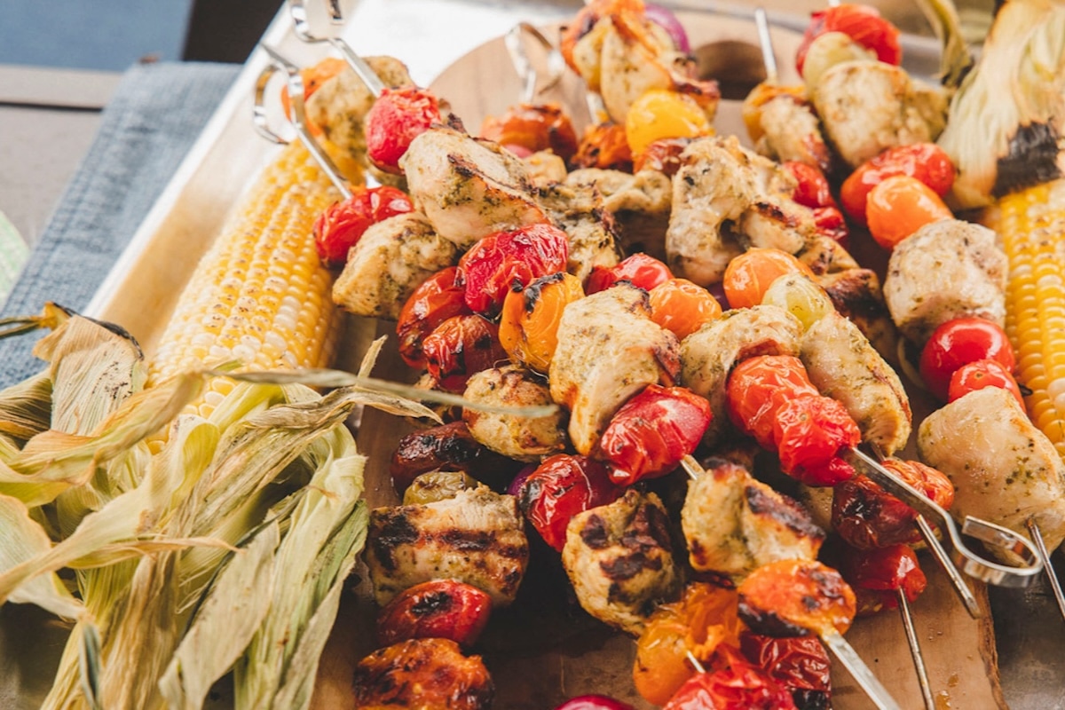 Grilled chicken and vegetable skewers with roasted cherry tomatoes are served on a platter next to ears of corn with husks, creating a colorful and appetizing summer meal. GREENHOUSE Goodness