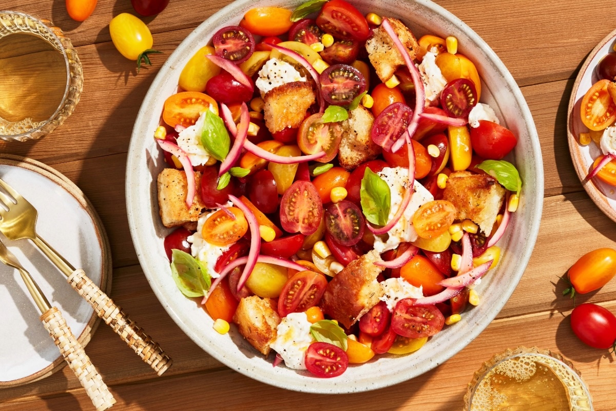 A bowl of colorful tomato panzanella salad with cherry tomatoes, toasted bread cubes, mozzarella, red onion, corn, fresh basil, and cracked pepper on a wooden table, beside drinks and utensils. GREENHOUSE Goodness