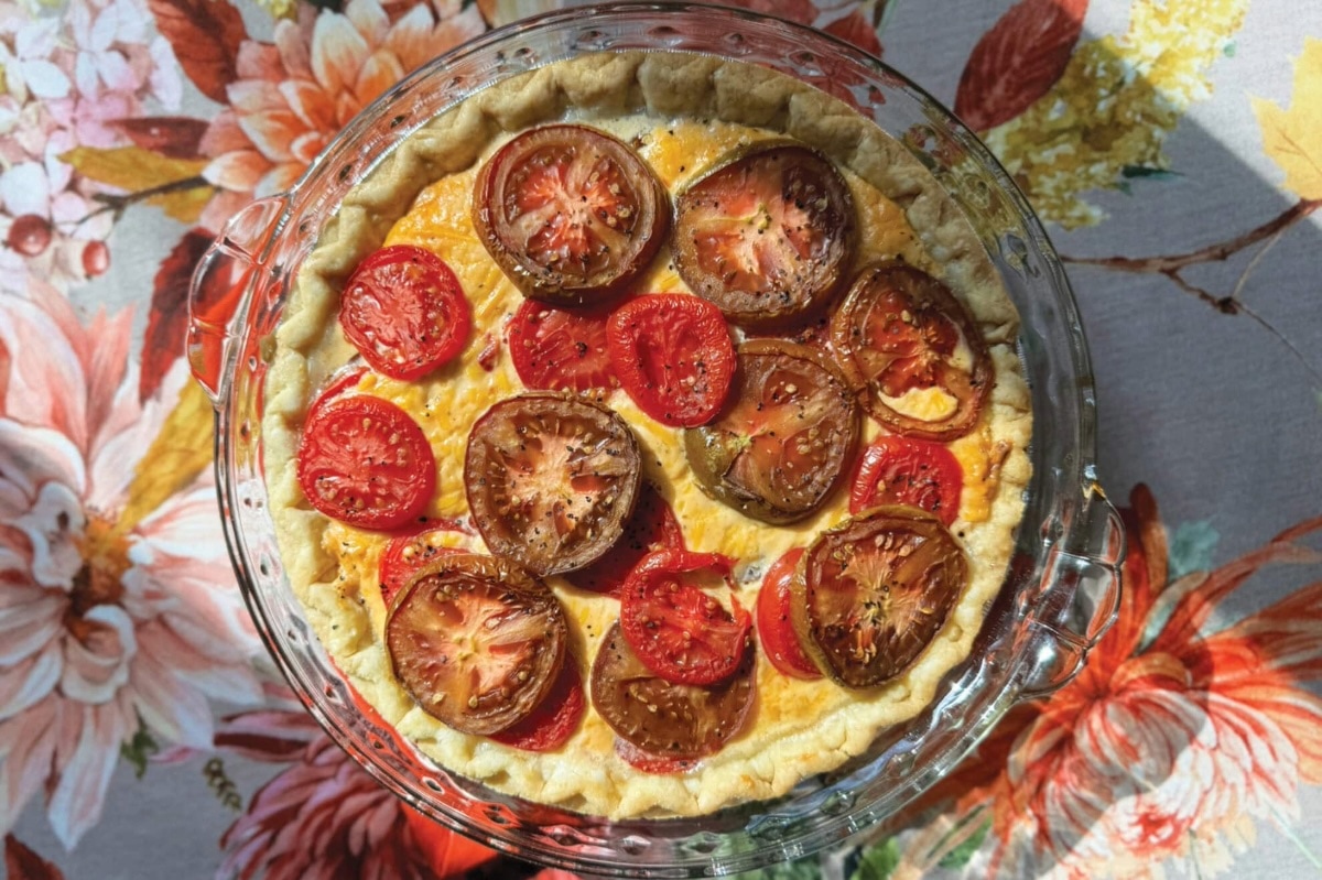 A baked tomato pie with a golden crust, topped with sliced red and brown tomatoes, sits in a glass pie dish on a floral-patterned tablecloth. GREENHOUSE Goodness