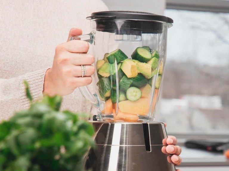 A person holding a blender filled with fresh cucumber, pineapple, and melon pieces, preparing to blend them in a modern kitchen. GREENHOUSE Goodness