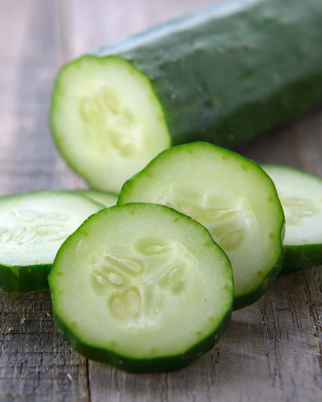 A close-up of a cucumber on a wooden surface, with three round slices in front of a larger uncut portion. The slices show the pale green interior and seeds. GREENHOUSE Goodness