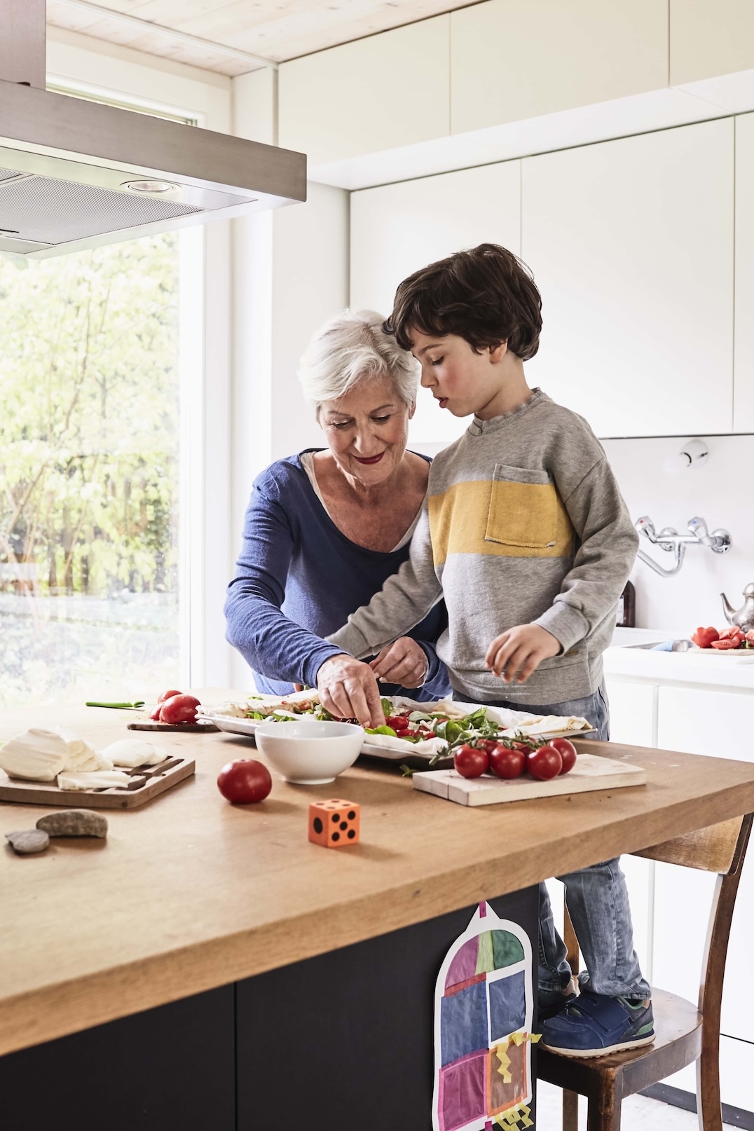 An older woman and a young boy prepare food together in a bright kitchen. The woman chops vegetables while the boy stands on a stool, helping her. Fresh ingredients and kitchen items are on the wooden countertop. GREENHOUSE Goodness