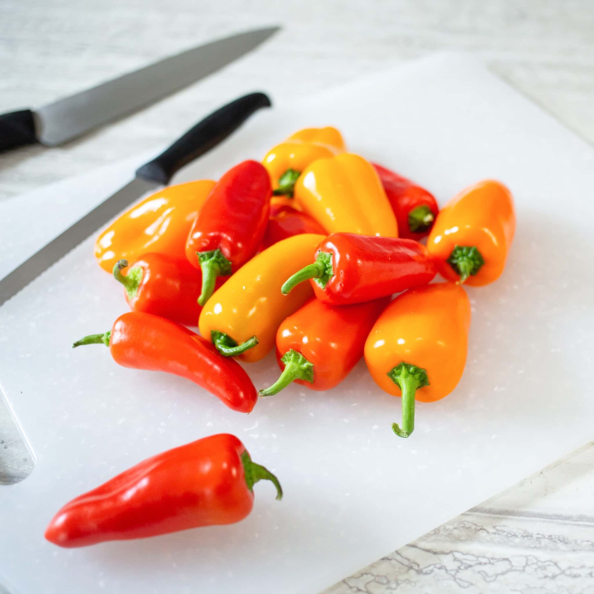 A pile of fresh red and orange mini sweet peppers on a white cutting board, with two kitchen knives placed nearby on a light-colored countertop. GREENHOUSE Goodness