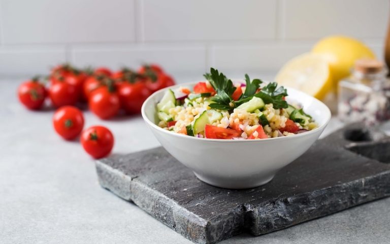 A white bowl filled with a fresh salad of couscous, cucumber, tomato, and parsley sits on a slate board. In the background, there are whole cherry tomatoes, a lemon, and kitchen items on a light countertop. GREENHOUSE Goodness