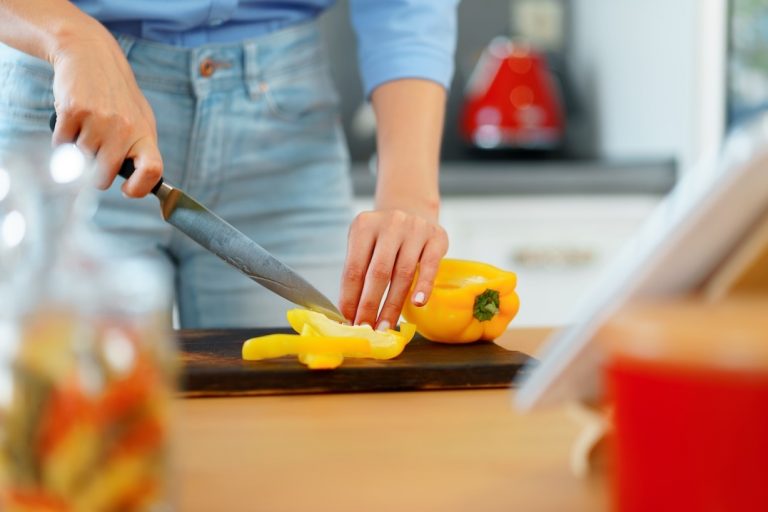 A person in jeans slices a yellow bell pepper on a wooden cutting board in a kitchen, with a red kettle and other kitchen items blurred in the background. GREENHOUSE Goodness