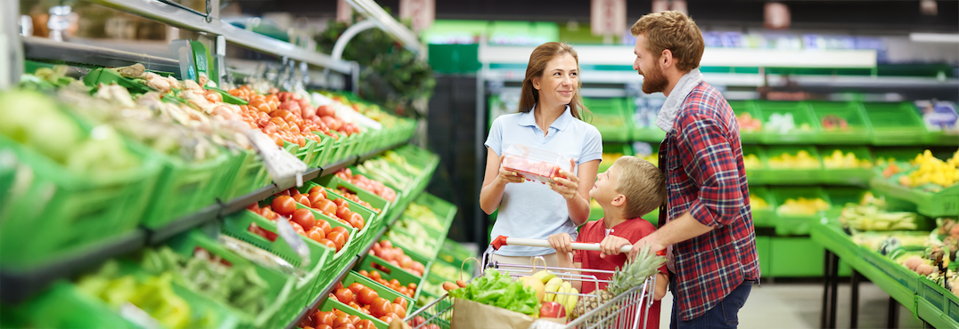 A smiling family with a shopping cart picks out fresh produce in the fruit and vegetable section, discussing greenhouse grown vs. field-grown options. The mother holds a food item while the father and young son look at her. GREENHOUSE Goodness