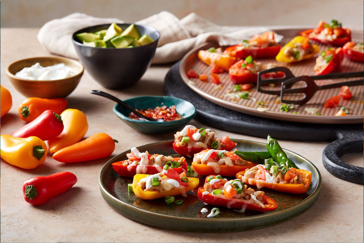A plate of colorful mini bell peppers stuffed with toppings like cheese, green onions, and diced tomatoes sits on a table, surrounded by bowls of avocado, sour cream, and red pepper flakes. More stuffed peppers are on a tray in the background. GREENHOUSE Goodness