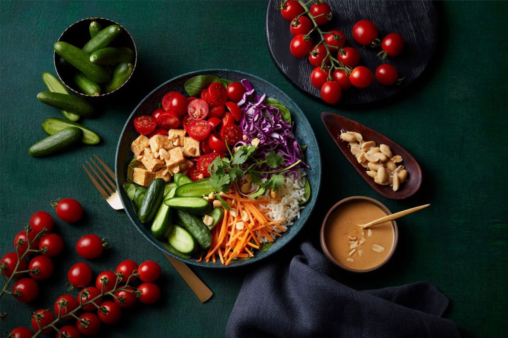 A colorful bowl with tofu, rice, cucumbers, cherry tomatoes, shredded carrots, and purple cabbage, garnished with cilantro. Surrounding are cherry tomatoes, cucumbers, peanuts, and a bowl of sauce on a dark green surface. GREENHOUSE Goodness