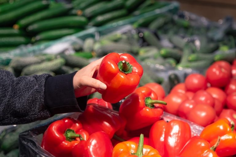 A person’s hand holds a large red bell pepper above a pile of other bell peppers in a grocery store, with cucumbers displayed in the background. GREENHOUSE Goodness