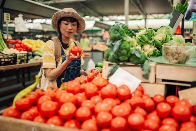 A woman wearing a hat and striped shirt examines greenhouse grown tomatoes at an outdoor market, surrounded by crates of fresh tomatoes, broccoli, and other vegetables. GREENHOUSE Goodness