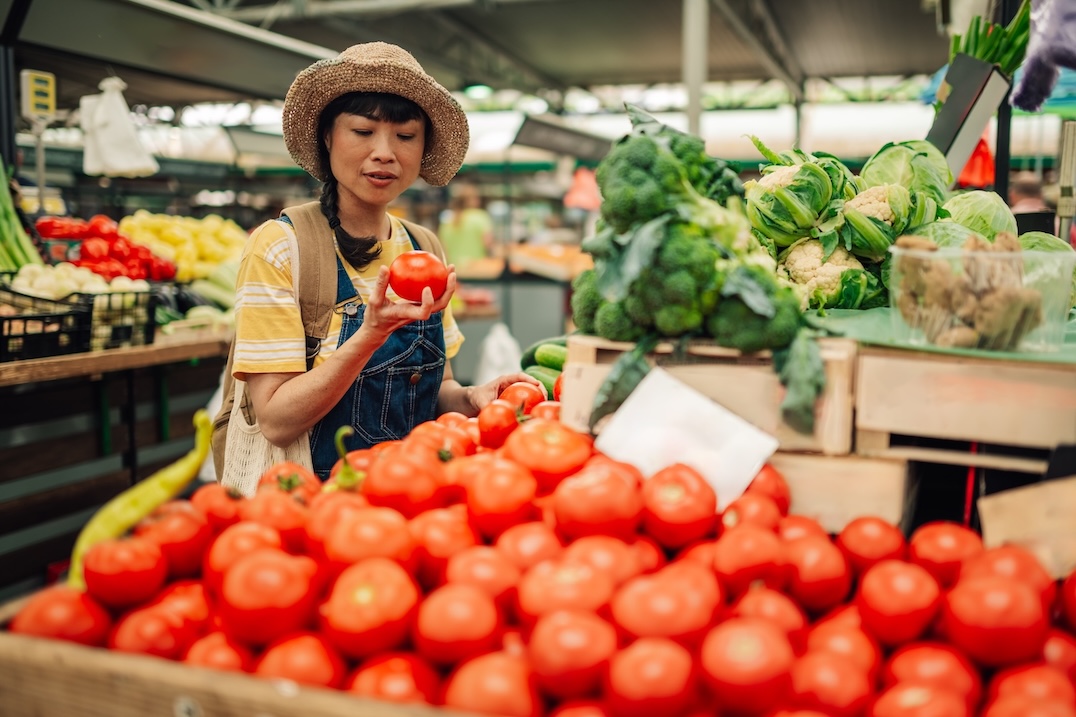 A woman wearing a hat and striped shirt examines greenhouse grown tomatoes at an outdoor market, surrounded by crates of fresh tomatoes, broccoli, and other vegetables. GREENHOUSE Goodness