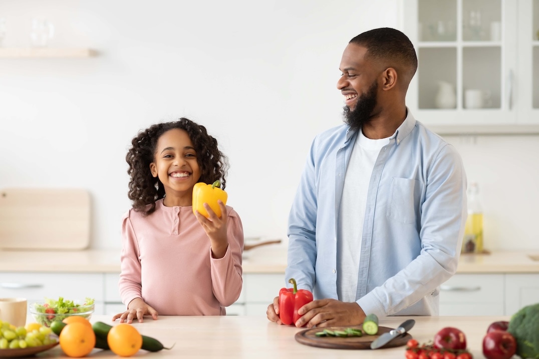 A smiling girl holds a yellow bell pepper while standing next to a man chopping vegetables in a bright kitchen; both appear happy and surrounded by fresh produce. GREENHOUSE Goodness