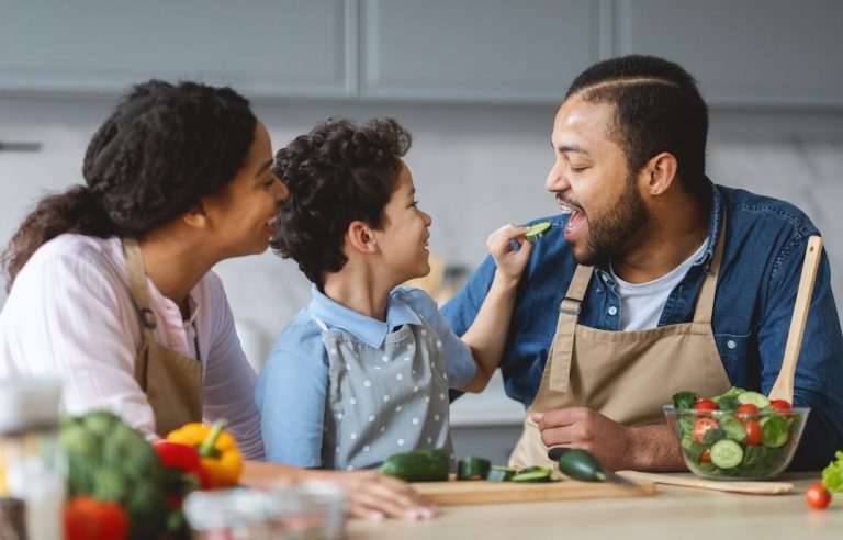 A smiling family in aprons prepares vegetables in a kitchen. A young boy playfully feeds a cucumber slice to an adult man, while a woman watches and smiles beside them. Fresh vegetables are on the table. GREENHOUSE Goodness