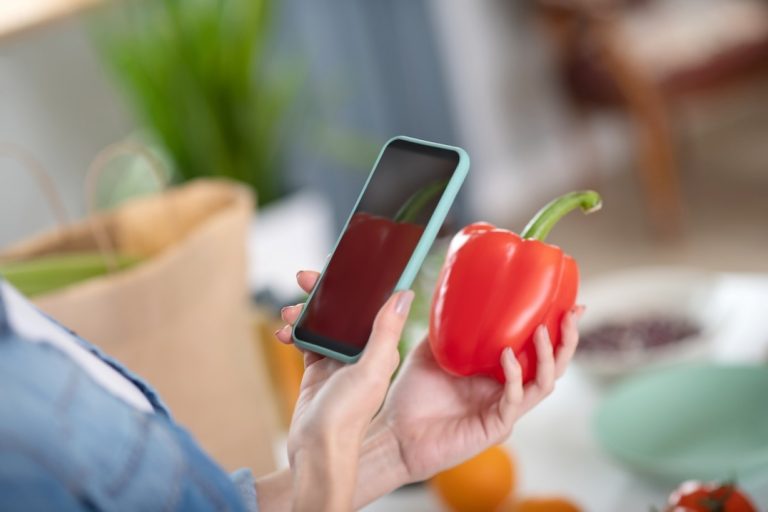 A person holds a red bell pepper in one hand and a smartphone in the other, possibly scanning or photographing the vegetable in a home kitchen setting. GREENHOUSE Goodness