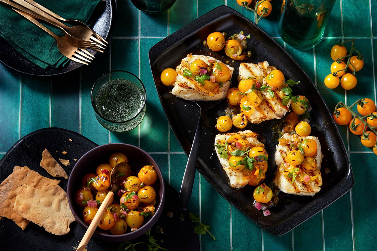 A black platter with grilled fish fillets topped with yellow cherry tomato salsa sits on a green-tiled table, surrounded by bowls of salsa, crisp flatbread, utensils, and glasses of sparkling drink. GREENHOUSE Goodness