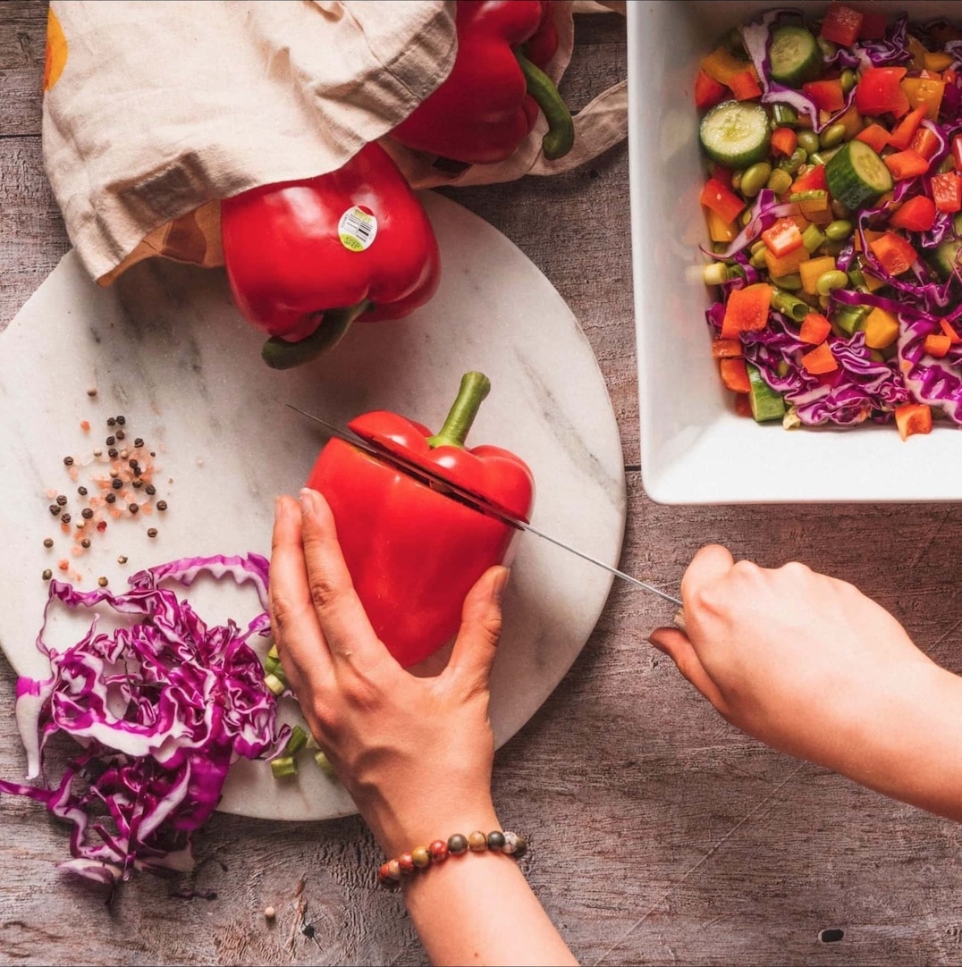 A person slices a red bell pepper on a marble cutting board surrounded by cabbage, whole peppers, peppercorns, and a bowl of fresh salad with cucumber, tomatoes, and cabbage on a wooden table. GREENHOUSE Goodness