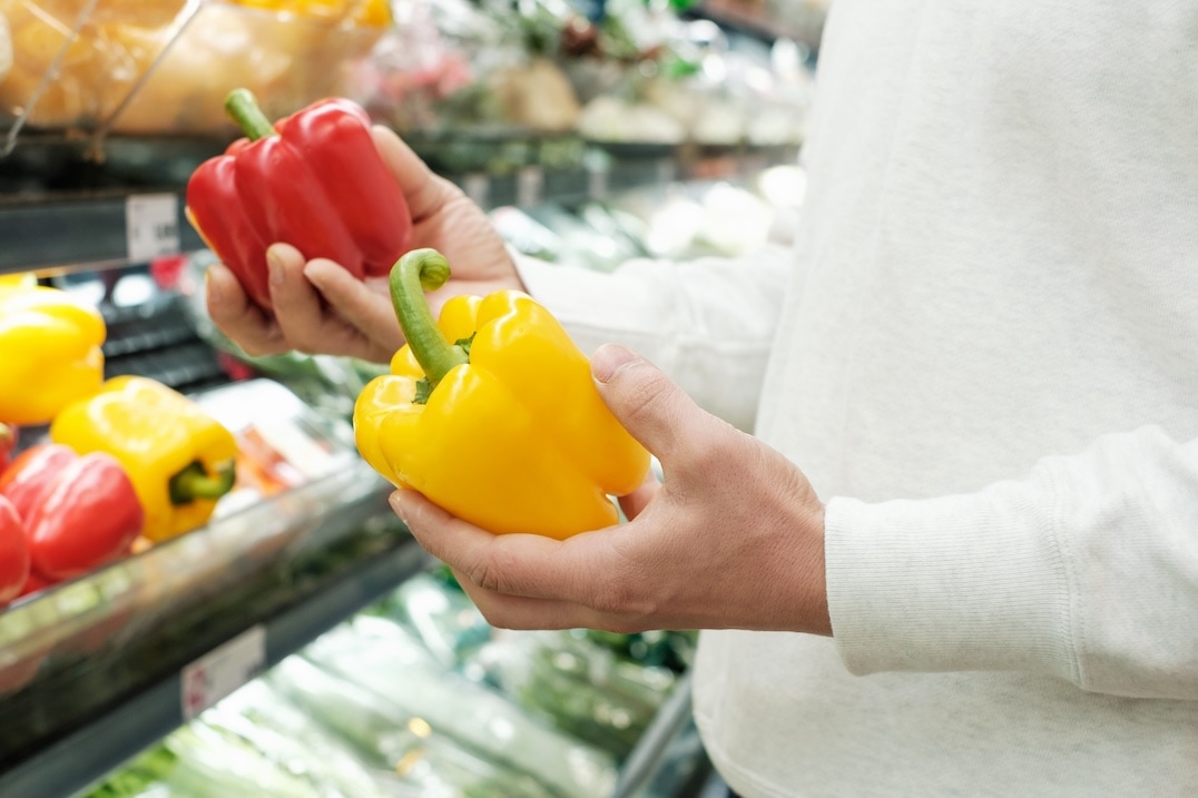 A person wearing a white sweater holds a red bell pepper in one hand and a yellow bell pepper in the other while shopping in the produce section of a grocery store. GREENHOUSE Goodness