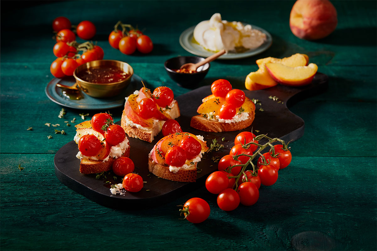 A dark board with toasted bread topped with ricotta cheese, roasted cherry tomatoes, and herbs. Fresh cherry tomatoes, peach slices, ricotta, and honey are arranged around on a teal wooden table. GREENHOUSE Goodness