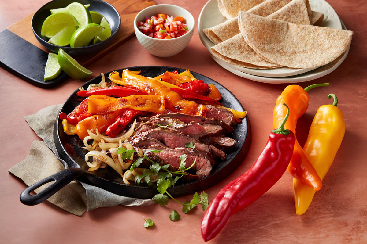 A skillet with sliced steak, grilled red and yellow peppers, and onions, garnished with cilantro. Nearby are lime wedges, salsa, whole wheat tortillas, and whole peppers on a peach-colored surface. GREENHOUSE Goodness