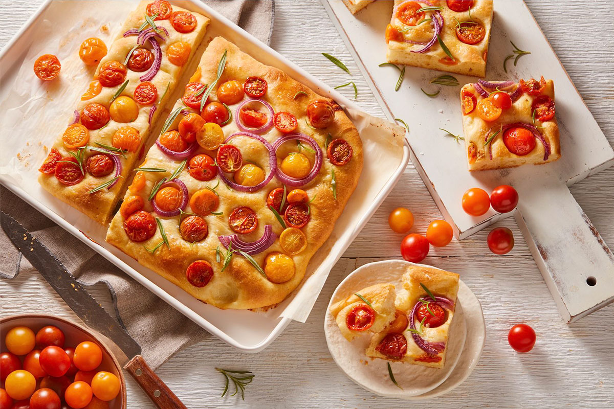 A tray of focaccia bread topped with cherry tomatoes, red onion, and rosemary, cut into squares. Some pieces are served on a plate and a white board, with fresh tomatoes scattered around. GREENHOUSE Goodness