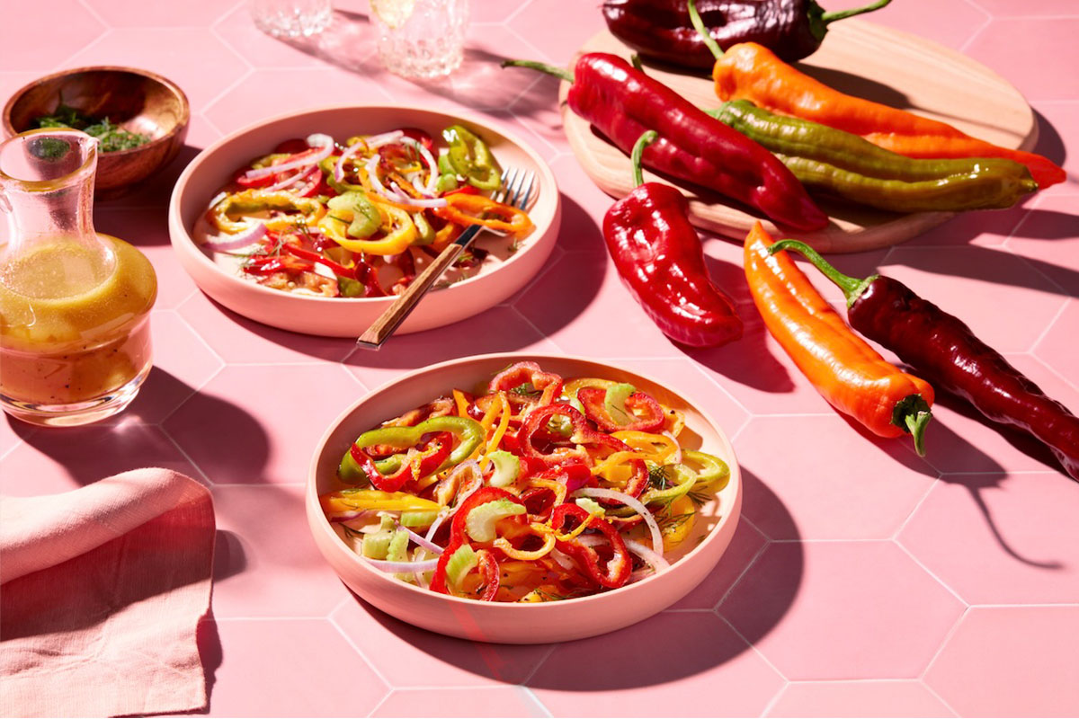 Two bowls of fresh salad with sliced bell peppers, onions, and celery are on a pink hexagon-tiled table, surrounded by whole colorful peppers, a small carafe of dressing, and a wooden board. GREENHOUSE Goodness