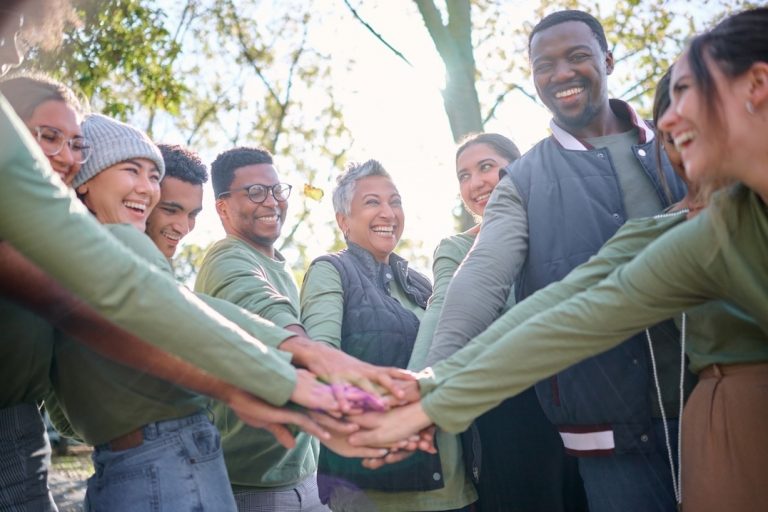 A diverse group of people stands in a circle outdoors, smiling and stacking their hands together in the center, showing teamwork and unity on a sunny day. GREENHOUSE Goodness