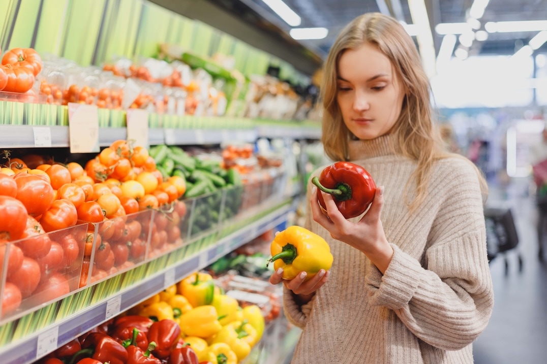 A woman in a beige sweater stands in a grocery store produce aisle, holding a red bell pepper and a yellow bell pepper, comparing them while surrounded by various fresh vegetables. GREENHOUSE Goodness