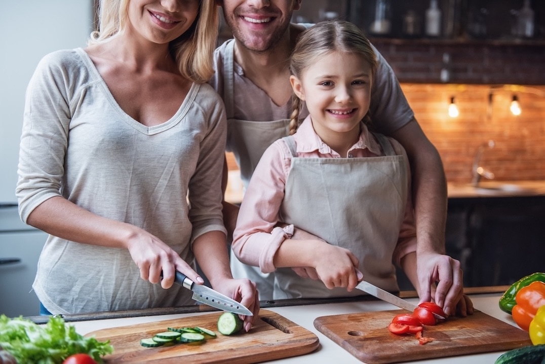 A smiling family of three, wearing aprons, prepares vegetables together in a kitchen. The woman slices cucumber, while the young girl proudly chops greenhouse peppers and tomato on a wooden board as the man stands behind them. GREENHOUSE Goodness