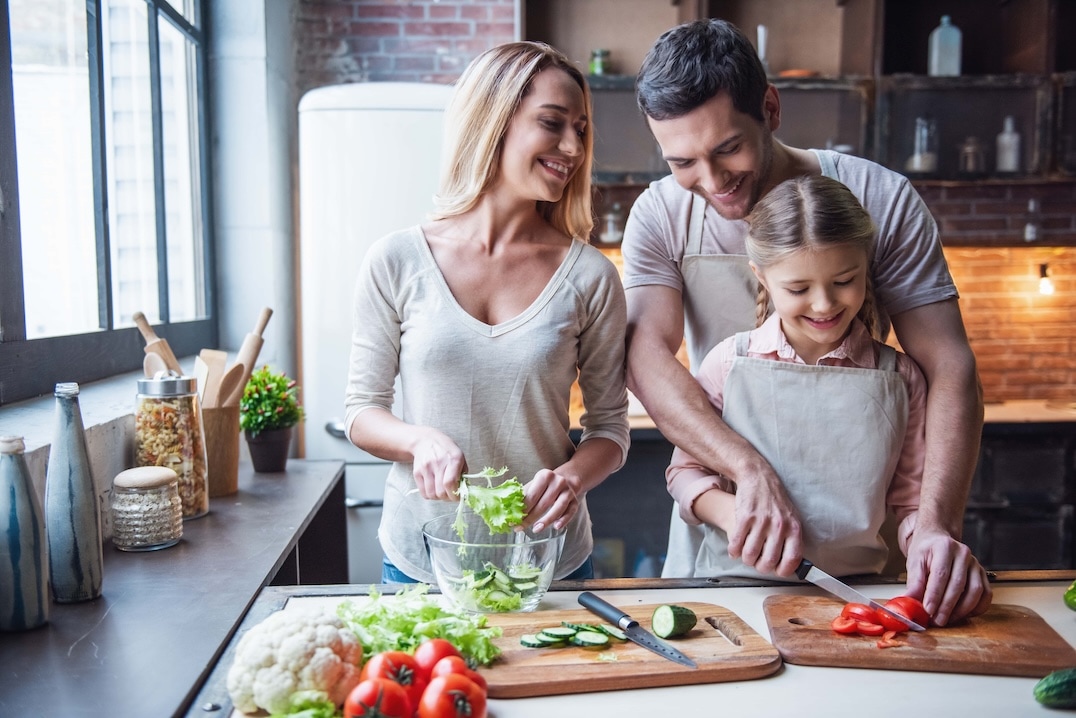 A smiling family of three prepares vegetables together in a kitchen. The woman mixes salad, the man slices a cucumber, and a young girl cuts a red bell pepper, all wearing casual clothes and aprons. GREENHOUSE Goodness