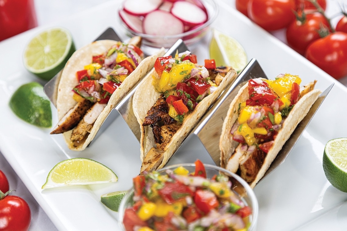 Three grilled chicken tacos topped with colorful mango salsa, served in metal taco holders on a white plate with lime wedges, a bowl of salsa, fresh radishes, and cherry tomatoes. GREENHOUSE Goodness