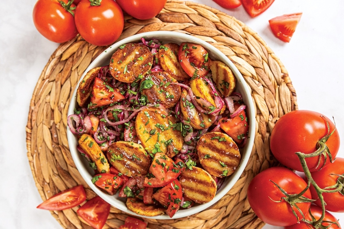 A bowl of grilled potato and tomato salad with red onions and chopped herbs, set on a woven placemat. Whole and sliced tomatoes surround the bowl on a light surface. GREENHOUSE Goodness