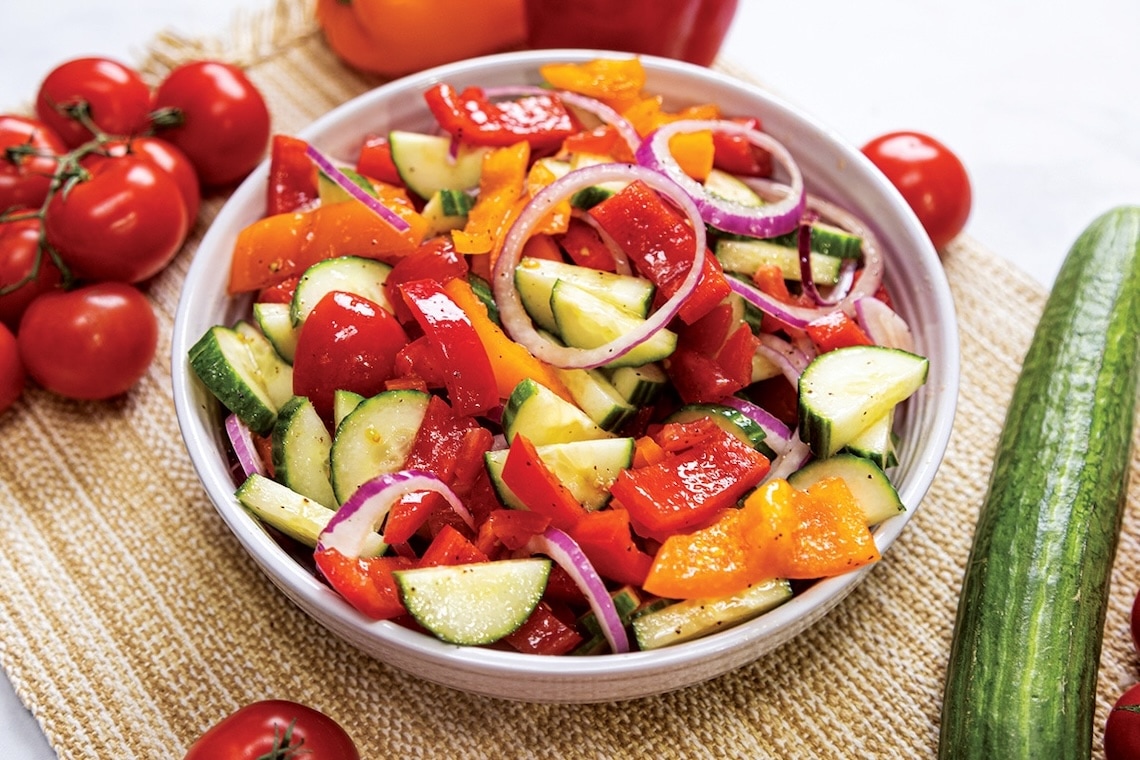 A bowl of fresh salad with chopped cucumbers, red and yellow bell peppers, red onions, and tomatoes, surrounded by whole tomatoes and a cucumber on a woven placemat. GREENHOUSE Goodness