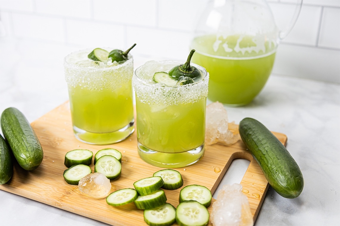 Two glasses of green cocktail with salted rims, garnished with sliced cucumber and a pepper, sit on a wooden board with ice cubes, whole and sliced cucumbers, and a pitcher of the same drink in the background. GREENHOUSE Goodness