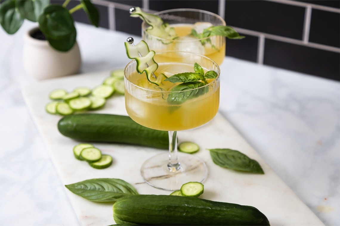 A cocktail garnished with basil and a cucumber ribbon sits on a marble surface, surrounded by cucumbers, cucumber slices, and basil leaves. Another drink and a potted plant are in the background. GREENHOUSE Goodness