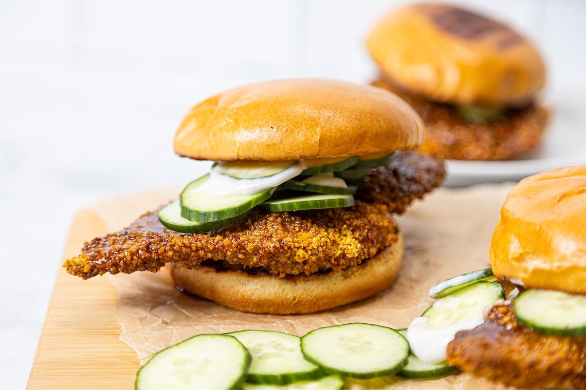 A crispy fried chicken sandwich on a brioche bun is topped with sliced cucumbers and sauce, with more cucumber slices and sandwiches visible in the background on a wooden surface. GREENHOUSE Goodness