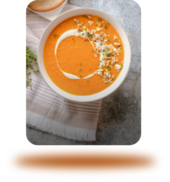 A bowl of creamy orange soup with a swirl of cream and sprinkled herbs, served on a striped napkin with a slice of bread in the background. GREENHOUSE Goodness