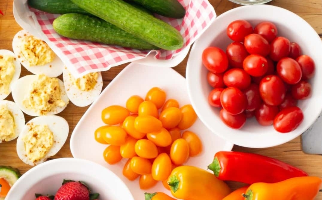 An assortment of fresh year-round greenhouse vegetables and deviled eggs, including cucumbers, red and yellow grape tomatoes, and mini bell peppers, arranged on a wooden surface in white dishes. GREENHOUSE Goodness