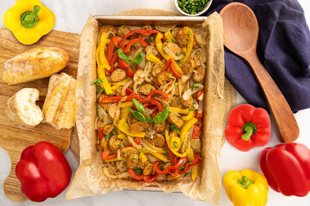 A baking tray lined with parchment paper holds a colorful dish of sliced sausages, red and yellow bell peppers, and onions. Surrounding the tray are fresh bell peppers, baguette pieces, chopped herbs, a wooden spoon, and a navy napkin. GREENHOUSE Goodness