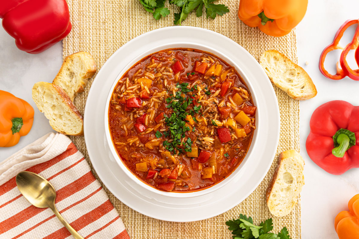 A bowl of hearty soup with rice, ground meat, and colorful bell peppers sits on a white plate, surrounded by baguette slices, whole bell peppers, parsley, a gold spoon, and a striped napkin on a woven placemat. GREENHOUSE Goodness